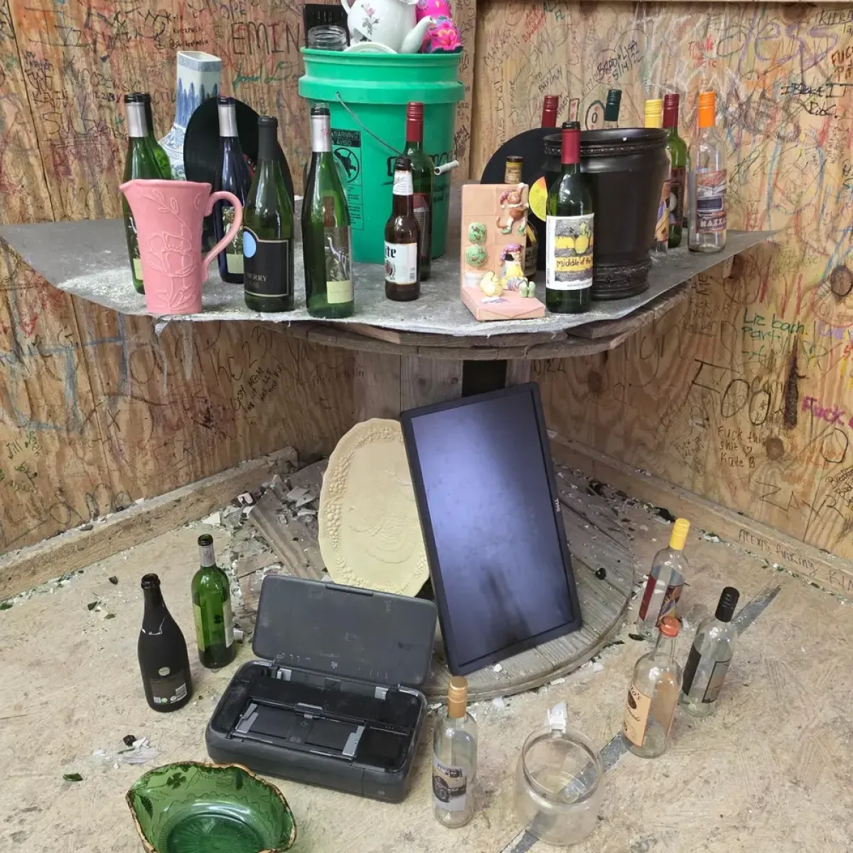 A cluttered corner with bottles, a green bucket, and an overturned monitor.