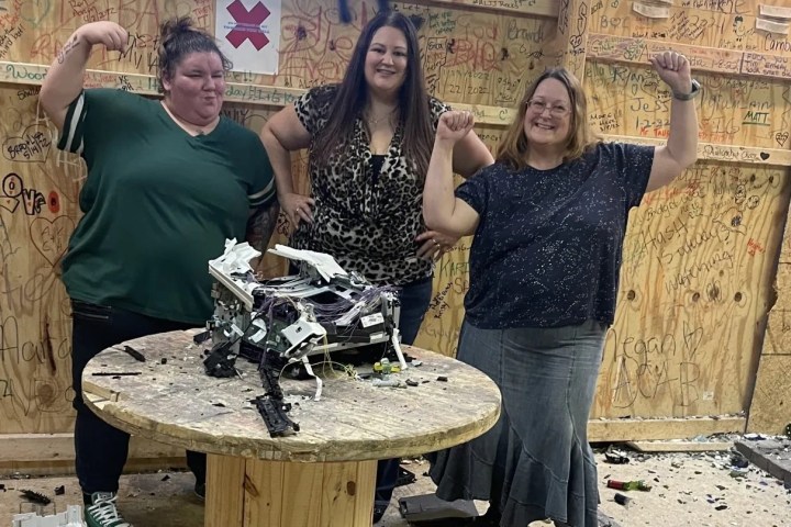 Three people posing triumphantly by a smashed item on a wooden table in a graffiti-covered room.