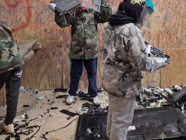 People in camo jackets and face shields in a room with broken electronics and graffiti-covered walls.
