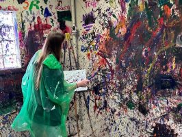 Person painting in a colorful room with paint splatters on walls.