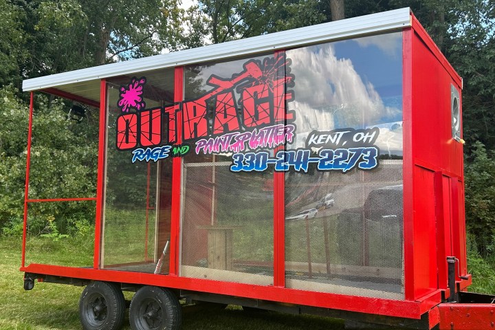 Red mobile paintball trailer with graphics and contact info, parked on grass.