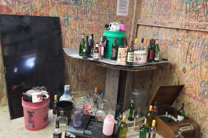 Room with graffiti walls, bottles on tables, and scattered items on the floor.