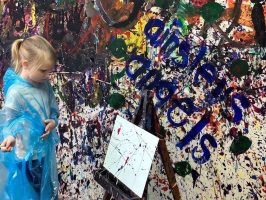 Child in blue poncho painting on canvas in colorful, splattered room.