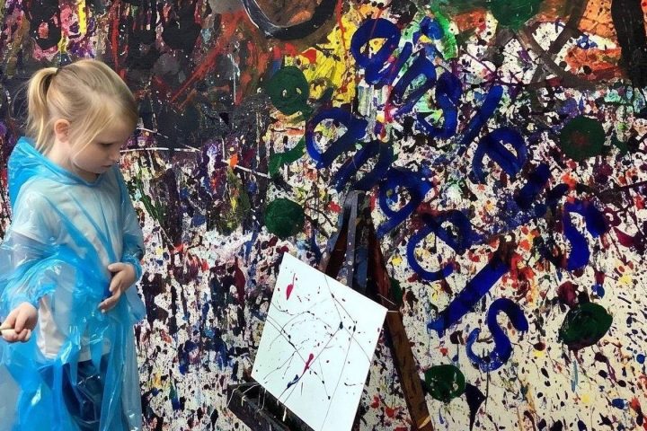 Child in blue poncho painting on canvas in colorful, splattered room.