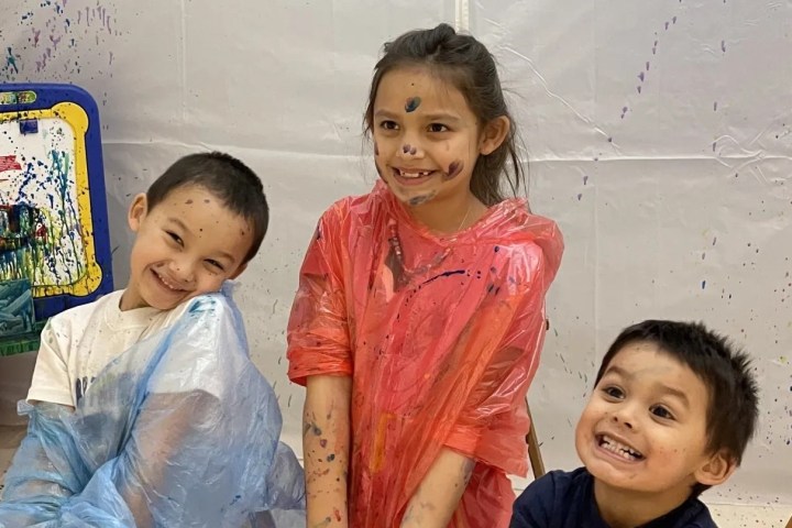 Three smiling children in paint-splattered ponchos with artwork in background.