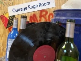 Vinyl record and bottles in a room with 'Outrage Rage Room' sign.